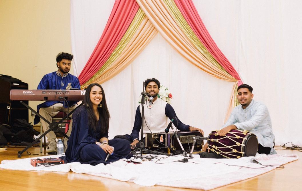 The group seated on stage in front of colourful drapes, with keyboard, harmonium, dholak and microphones set up