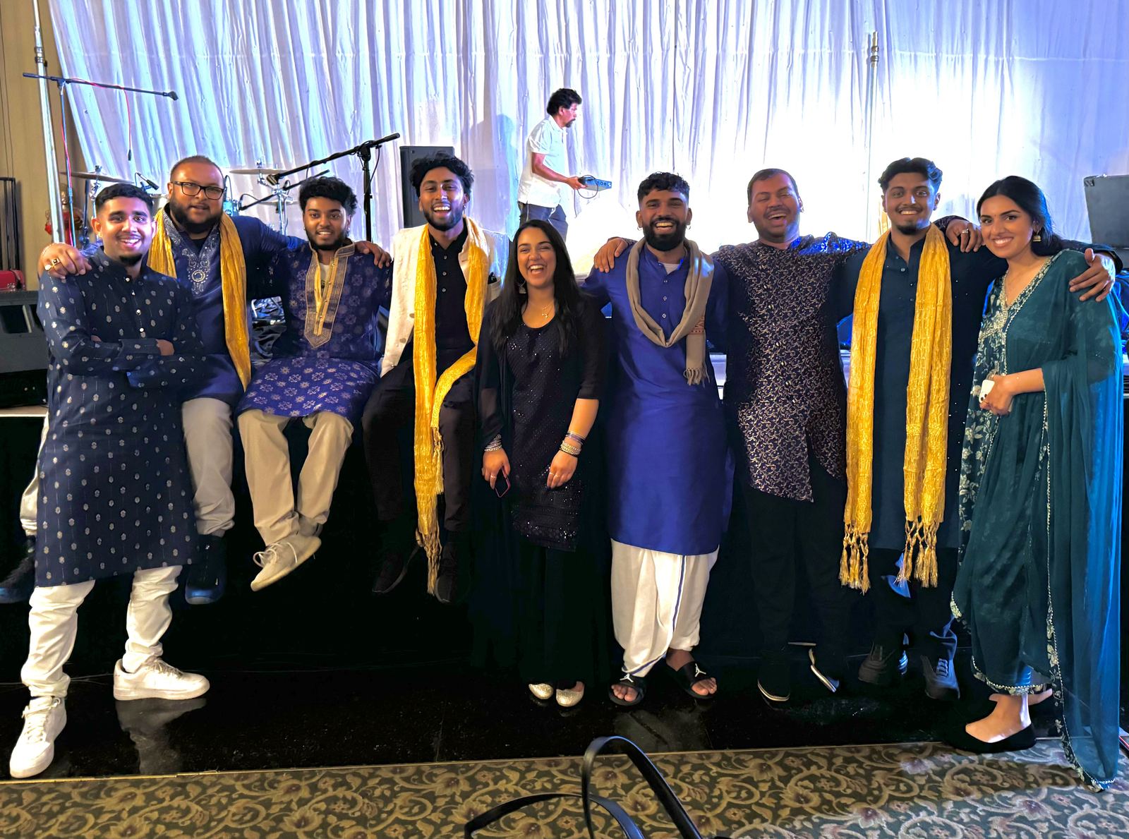 The full group in kurtas and scarves posing together on a ballroom stage